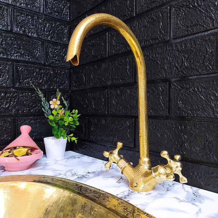 A hammered brass gooseneck faucet with cross handles, elegantly mounted on a marble countertop beside a matching brass sink, set against a striking black brick wall with decorative elements like a pink Moroccan tagine and a small potted plant.