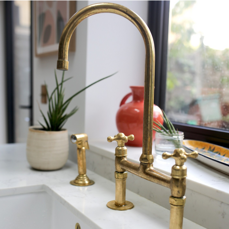 A brass bridge faucet with cross handles and a matching side sprayer, mounted on a white marble countertop beside a farmhouse sink, with warm decor and natural light.