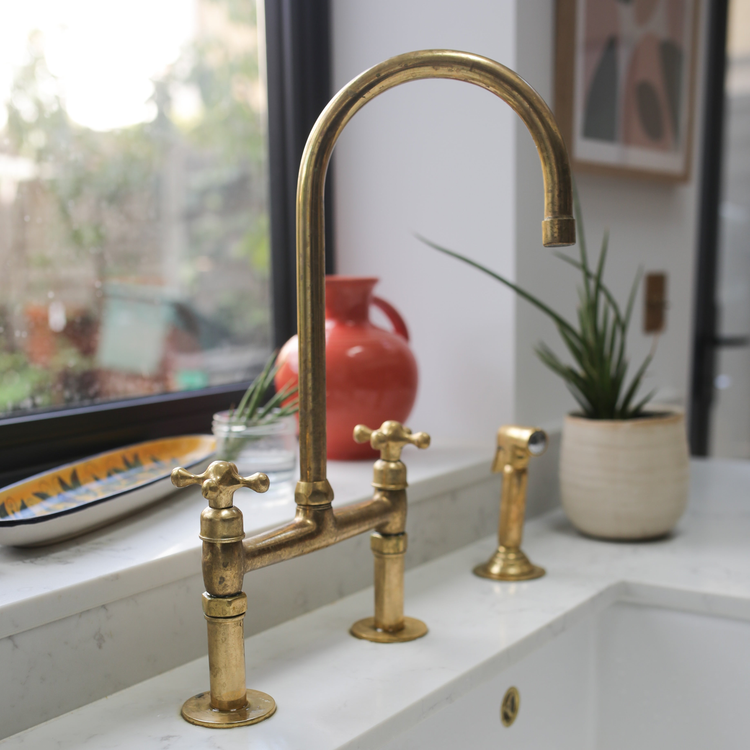 A brass bridge kitchen faucet with cross handles and a matching side sprayer, elegantly mounted on a farmhouse sink, surrounded by warm decor including a wooden countertop, fresh yellow flowers, and an open cookbook by the window.