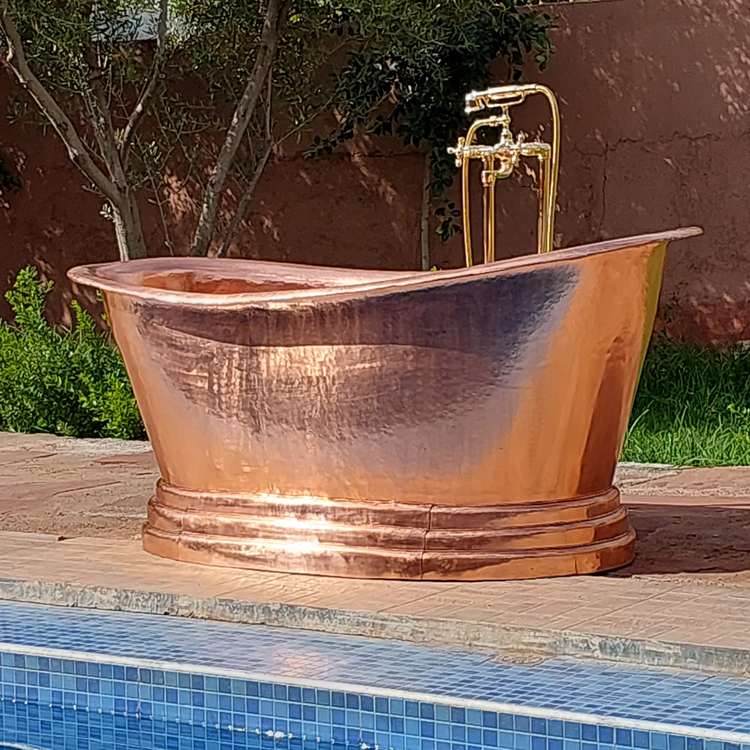 A freestanding hammered copper bathtub with an elegant curved rim and pedestal base, set outdoors near a pool, complemented by a brass floor-mounted faucet, creating a luxurious and timeless bathing experience.
