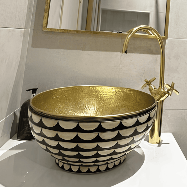 A luxurious brass vessel sink with a black and gold scalloped pattern, featuring a hammered interior for texture, paired with a tall brass gooseneck faucet with cross handles, set on a sleek white countertop with a gold-framed mirror above.