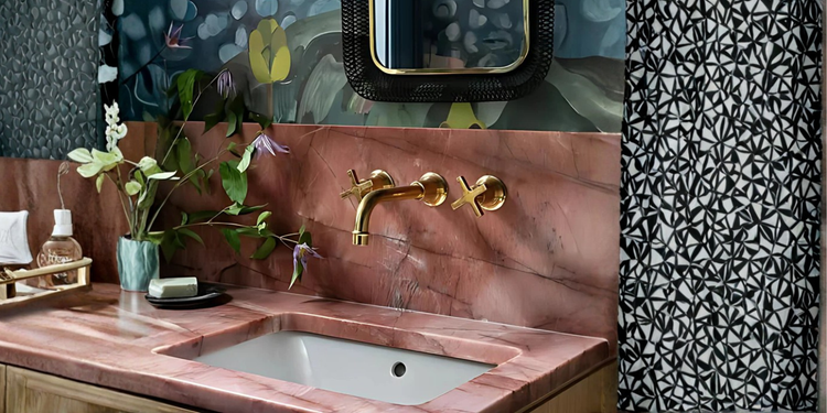 A vintage brass wall-mounted faucet with cross handles extending over a decorative ceramic basin with pink floral patterns, complemented by a mounted soap holder, a distressed mirror, and warm rustic accents.