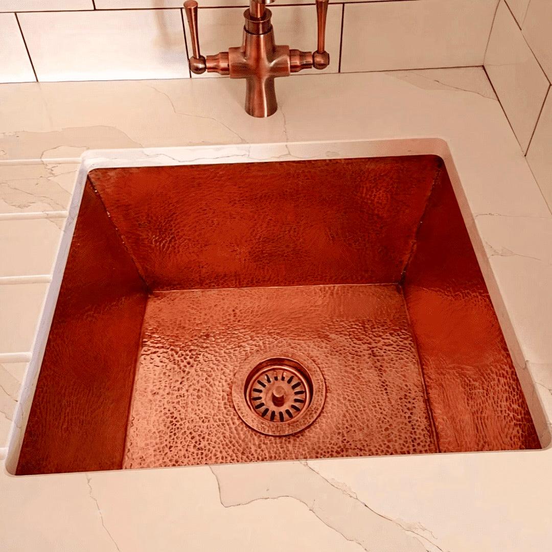 A hammered copper farmhouse sink with a rich, warm finish, seamlessly integrated into a white marble countertop, paired with a matching antique copper bridge faucet for a timeless and elegant kitchen design.
